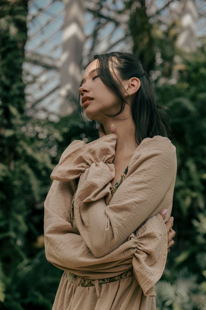A woman embraces herself in a beige boho dress amidst lush greenery inside a greenhouse.
